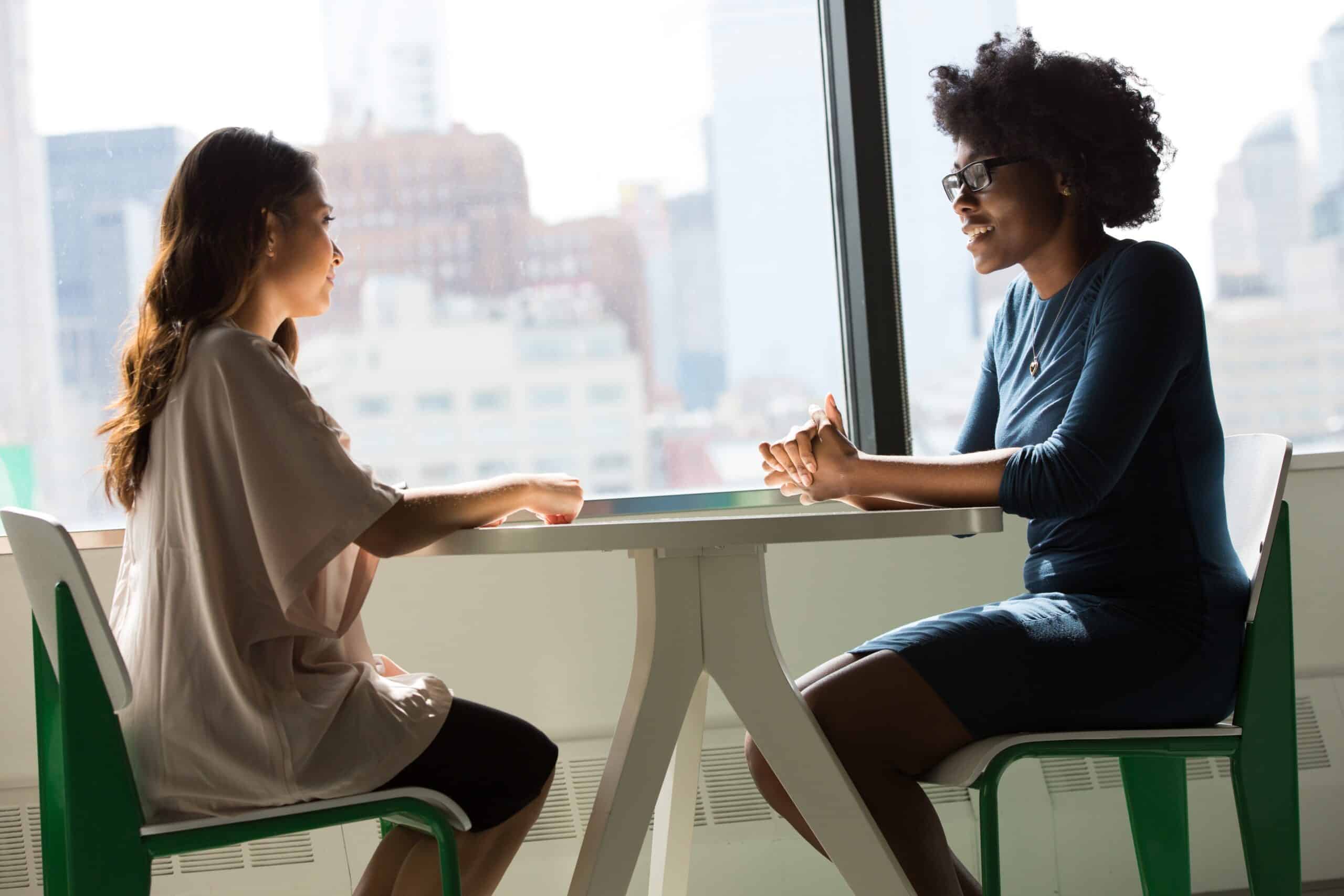 Two women sitting at a table, talking to one another in an corporate office